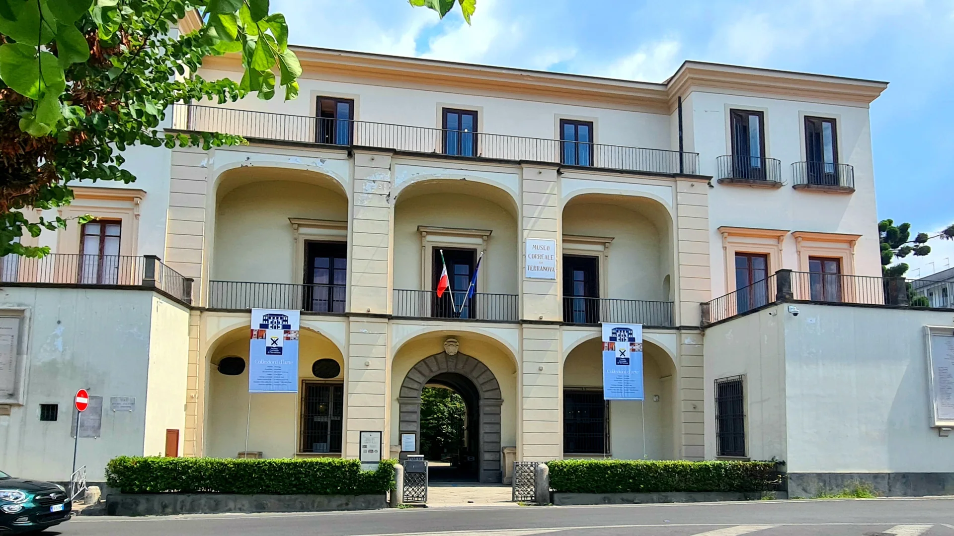 The cream-coloured front of the Museo Correale di Terranova in Sorrento, with three arched loggias above a stone gateway, iron-railed balconies, a tricolour flag at the entrance and a leafy branch in the corner.