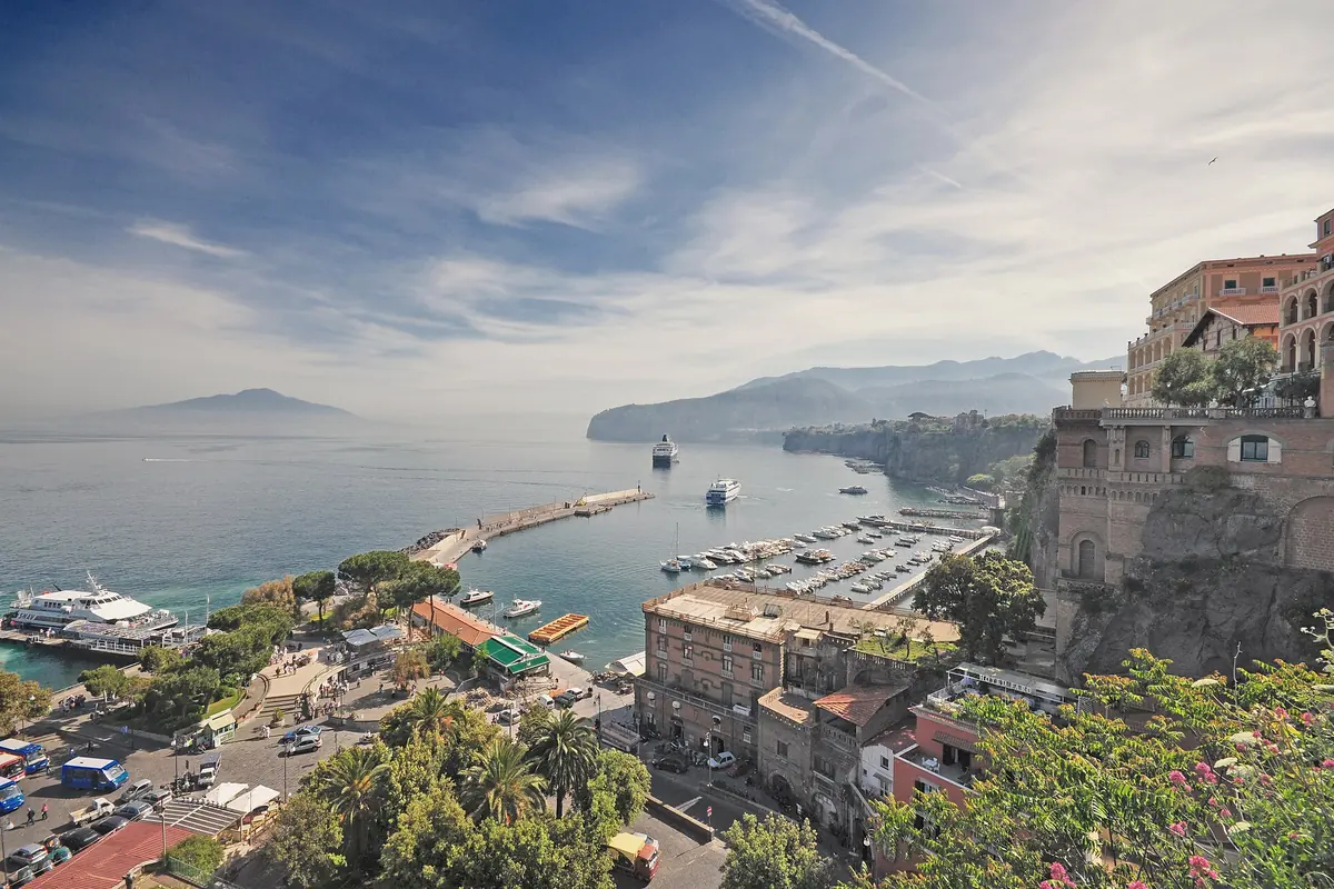 A view over the harbour of Sorrento with a long pier, small boats and cliffs on the right, and the dim shape of Vesuvius across the water under a streaked sky.