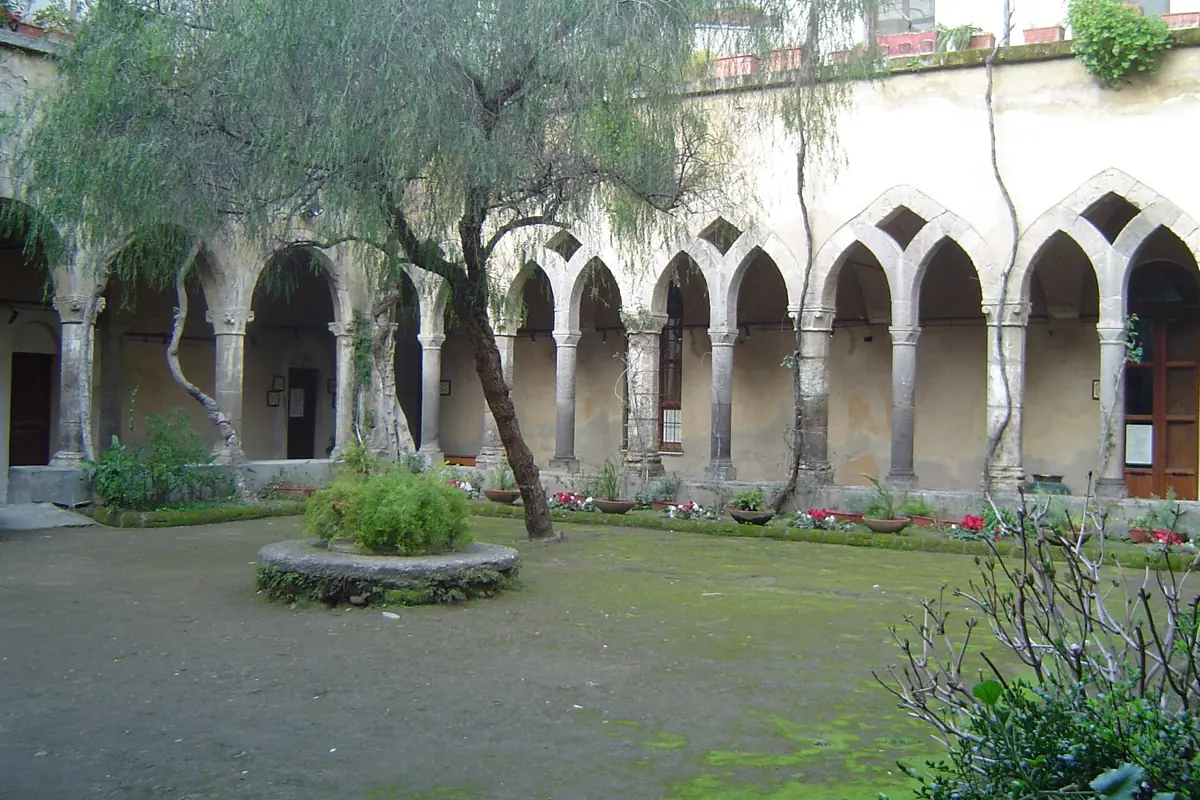A cloister garden in Sorrento with a line of pointed arches on slim columns and a round stone well in the grass.