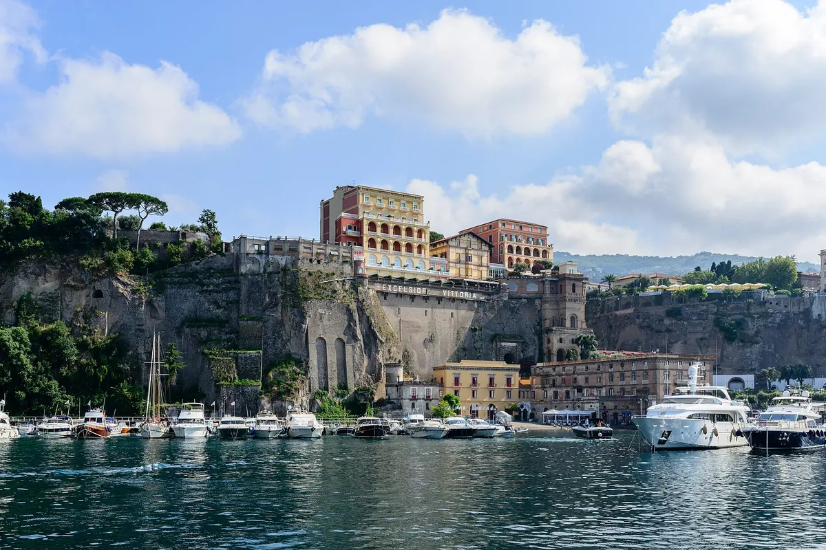 Moored boats and a white yacht in the harbour of Sorrento beneath a tall cliff crowned with pink and ochre buildings.