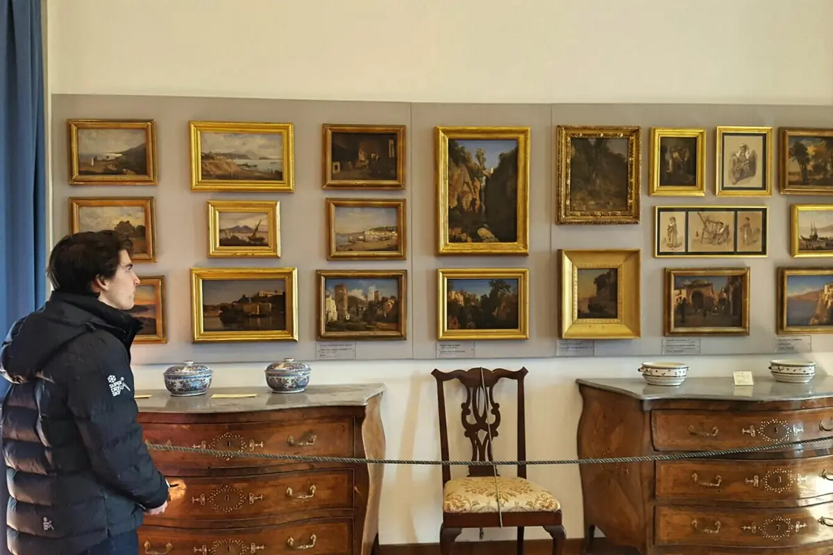 A museum room with a wall of small gilt-framed landscape paintings above two dark wooden chests of drawers and a chair.